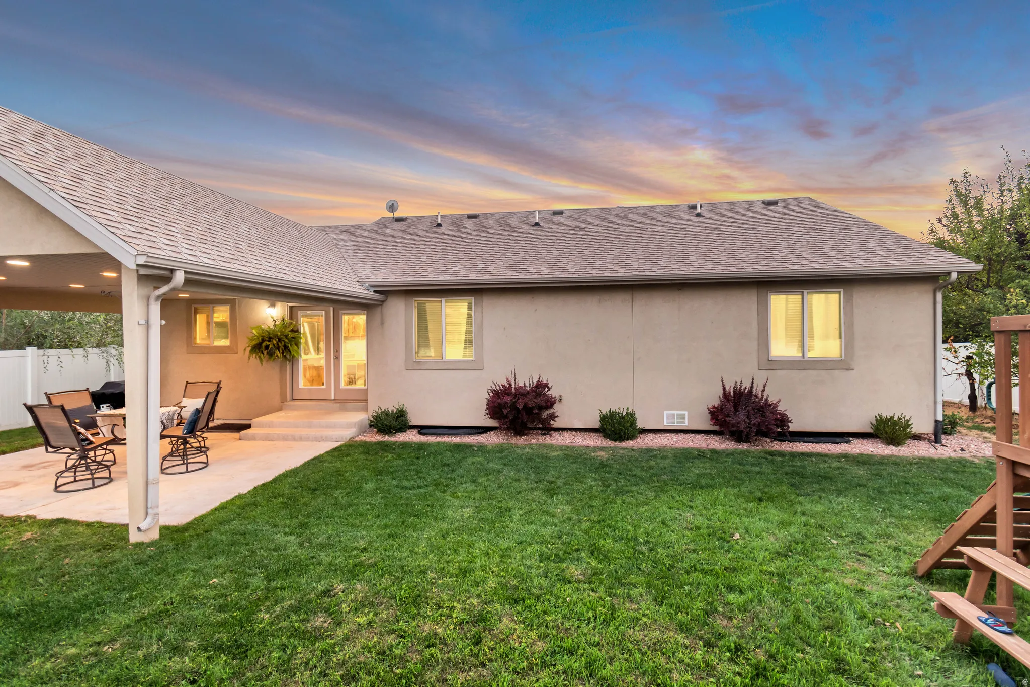 Expansive backyard featuring a manicured lawn, covered patio, stucco exterior, and white privacy fencing