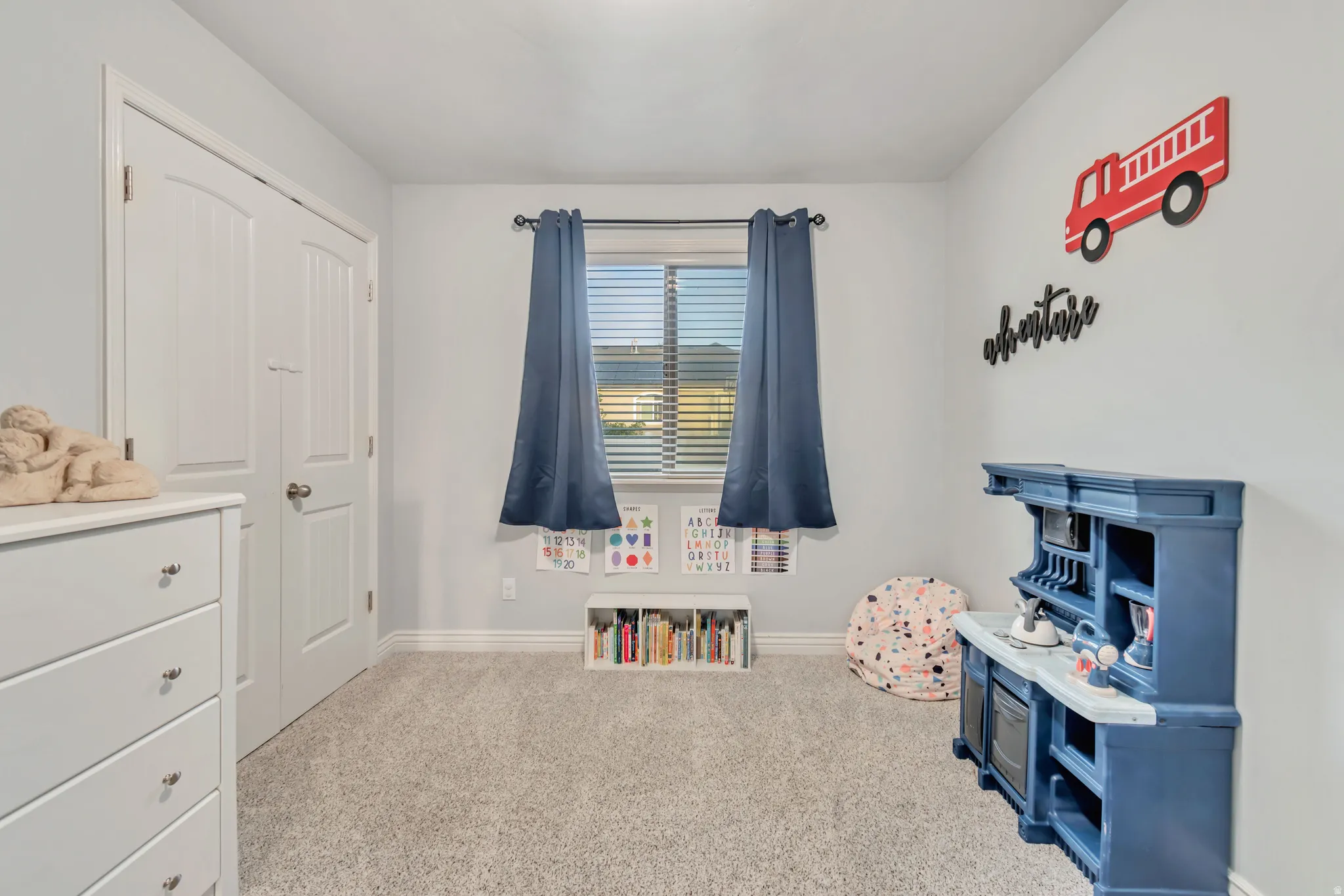 Room featuring light gray walls, gray carpet, a window with blinds and blue curtains, and white baseboards