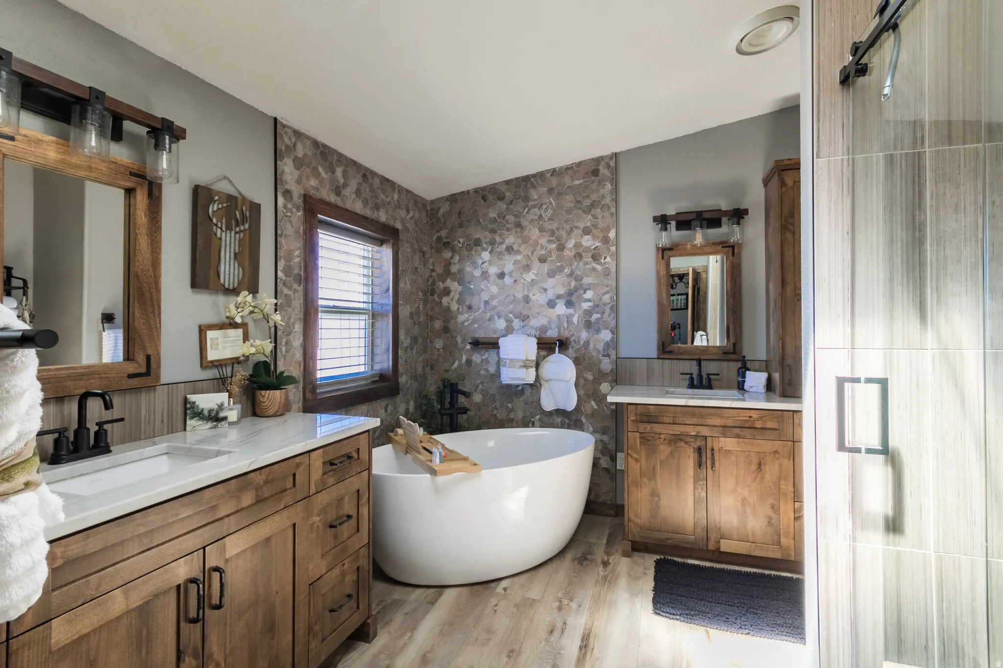 Bathroom featuring a freestanding soaking tub, dual wood-finish vanities with countertop sinks, a stone mosaic accent wall, and wood-finish flooring