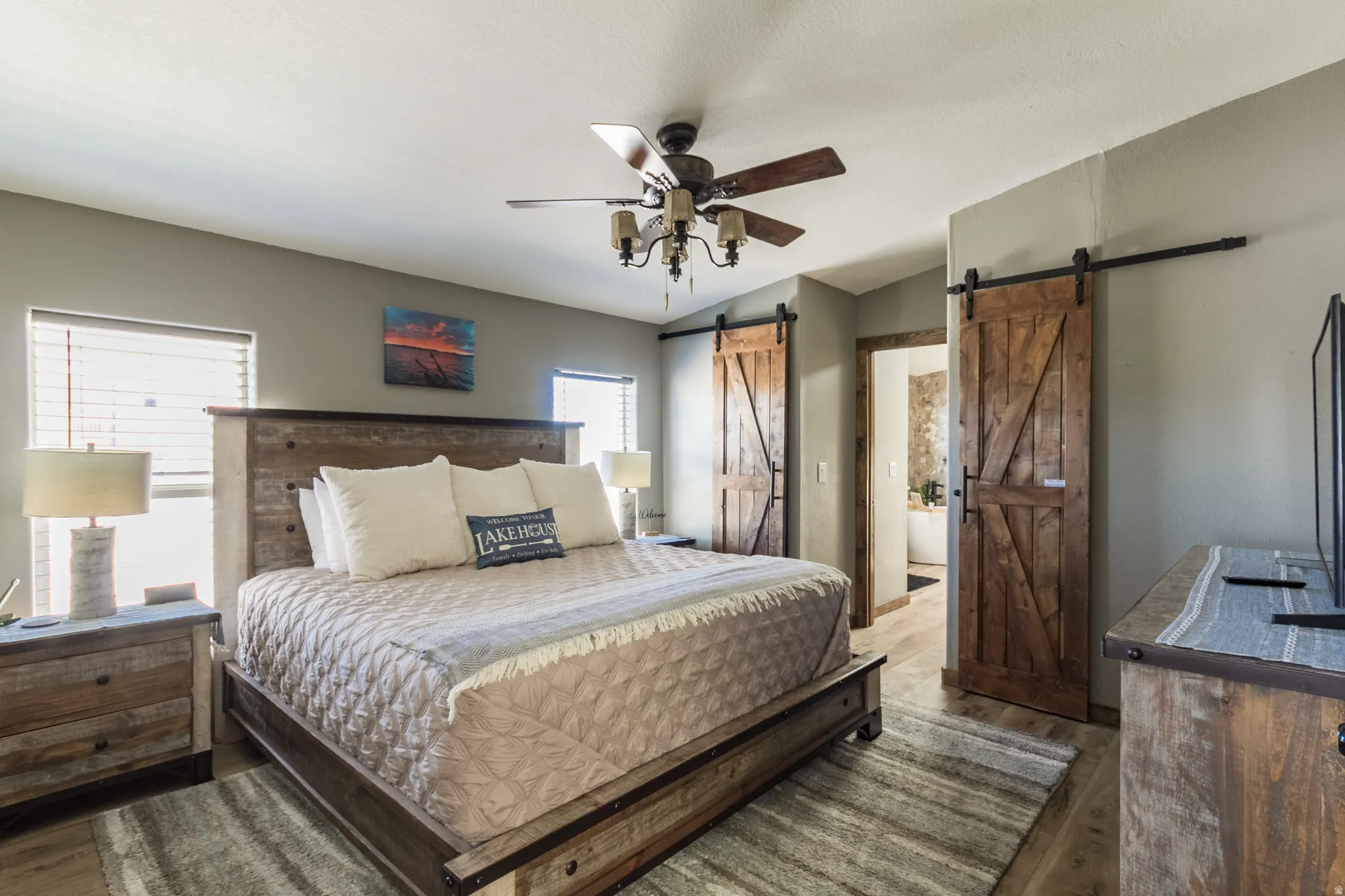Spacious room featuring wood-finish flooring, two large windows with blinds, a ceiling fan with light fixture, and two rustic wood barn doors