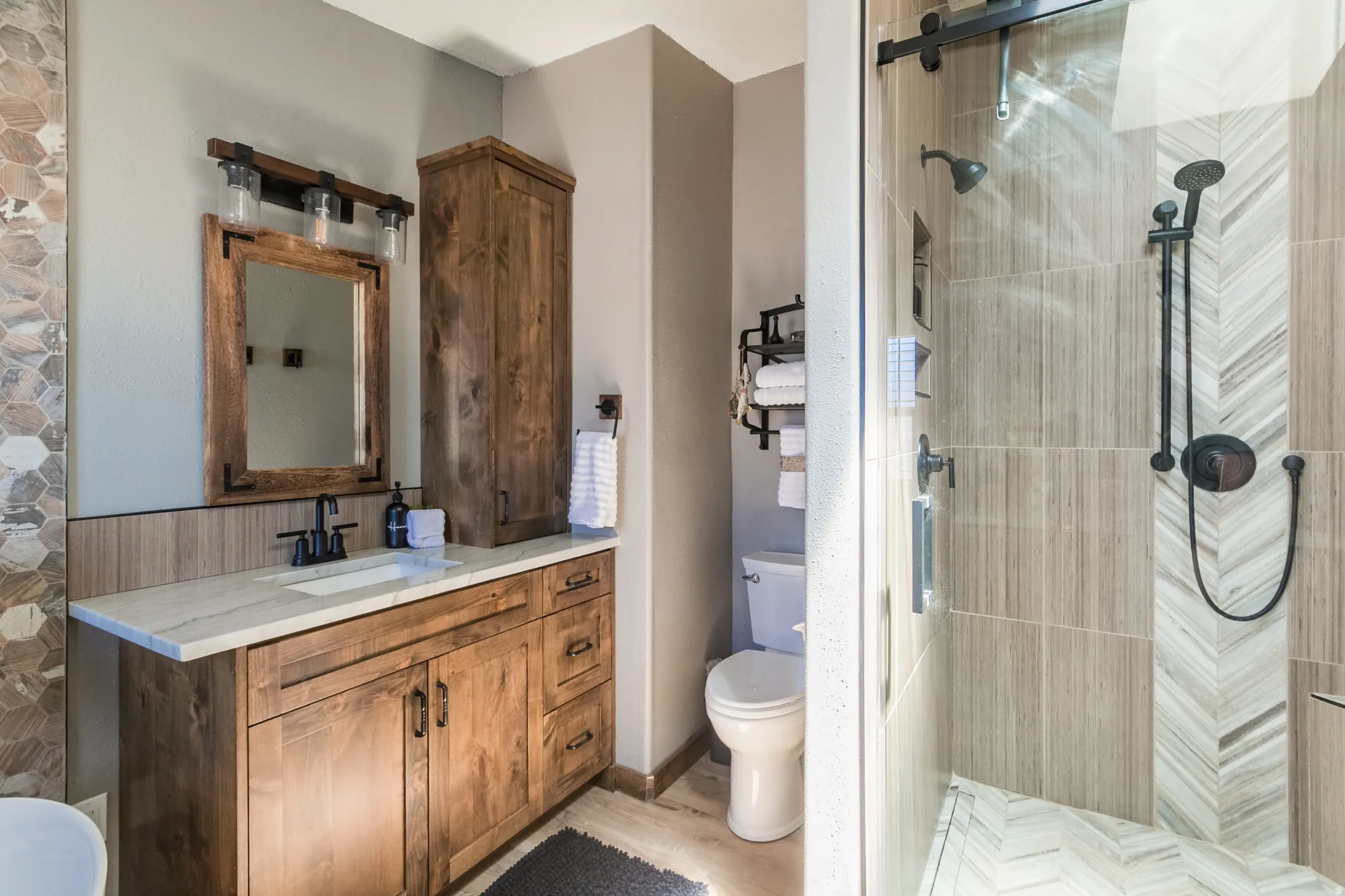 Bathroom featuring a wood-finish vanity with a light-toned countertop, a shower enclosure with a glass door, a wall-mounted showerhead, a separate handheld shower, and natural-toned tile flooring