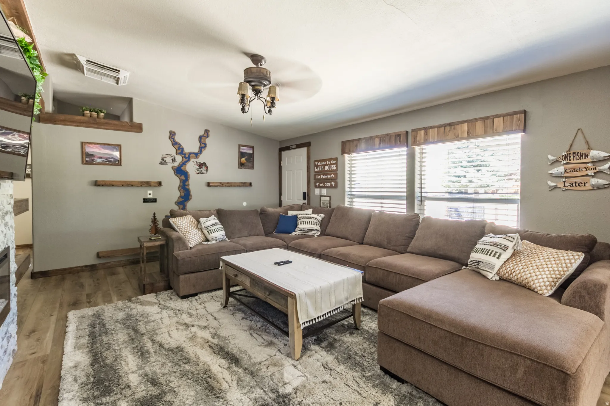 Spacious living area featuring wood-finish flooring, two large windows with horizontal blinds, and a ceiling fan with integrated lighting
