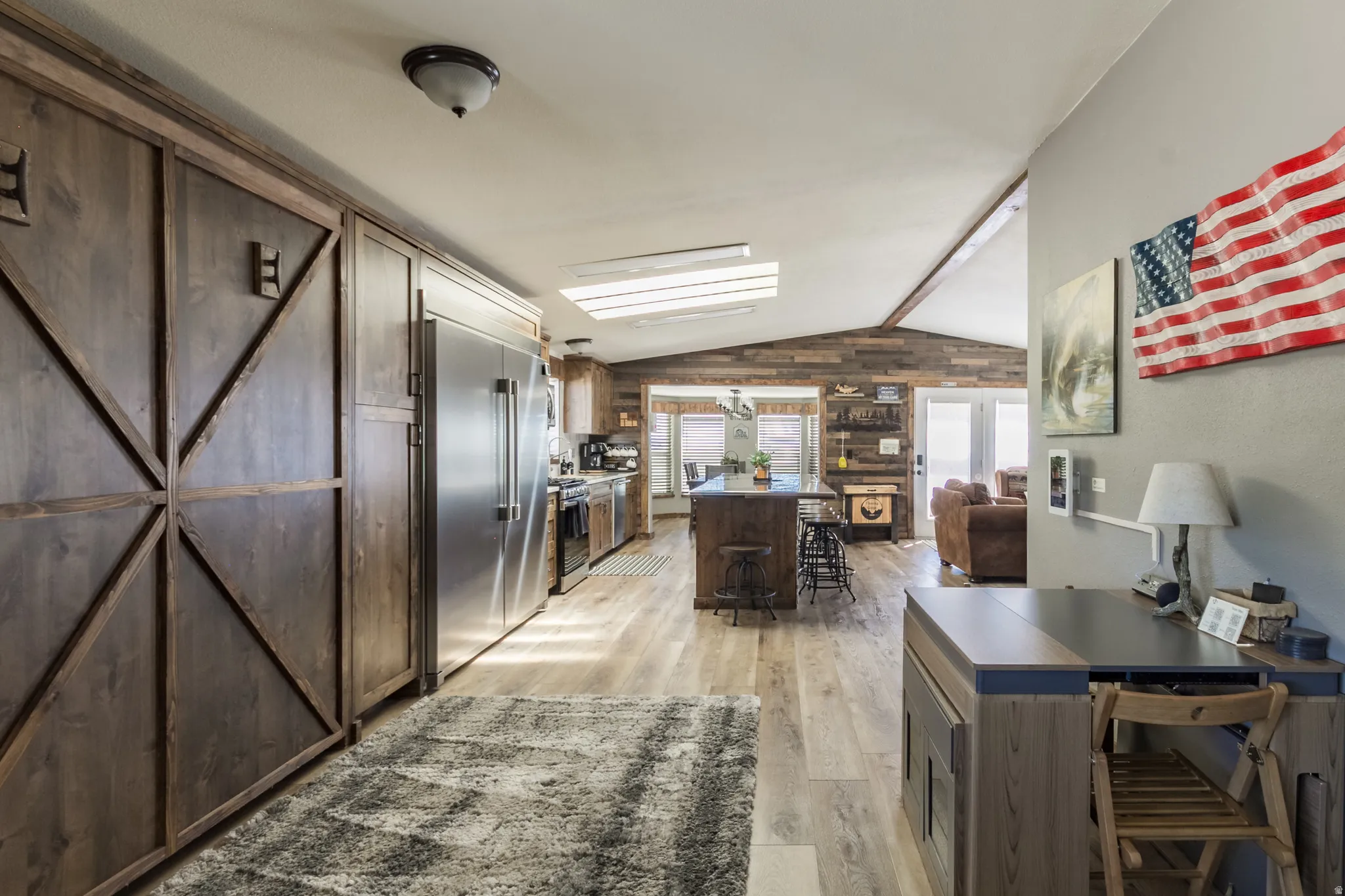Open-concept kitchen and living area featuring wood-finish flooring, vaulted ceilings with wood beams, and a wood-paneled accent wall