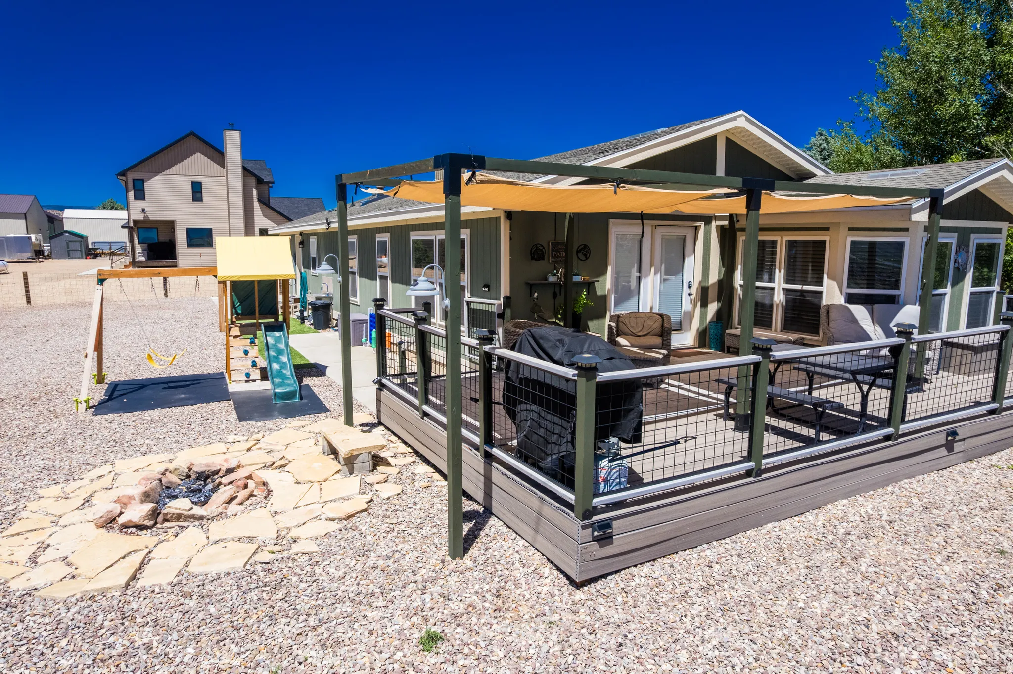 Expansive gravel yard featuring a composite deck with metal railing, a pergola with a fabric canopy, a stone fire pit, and a swing set with a slide