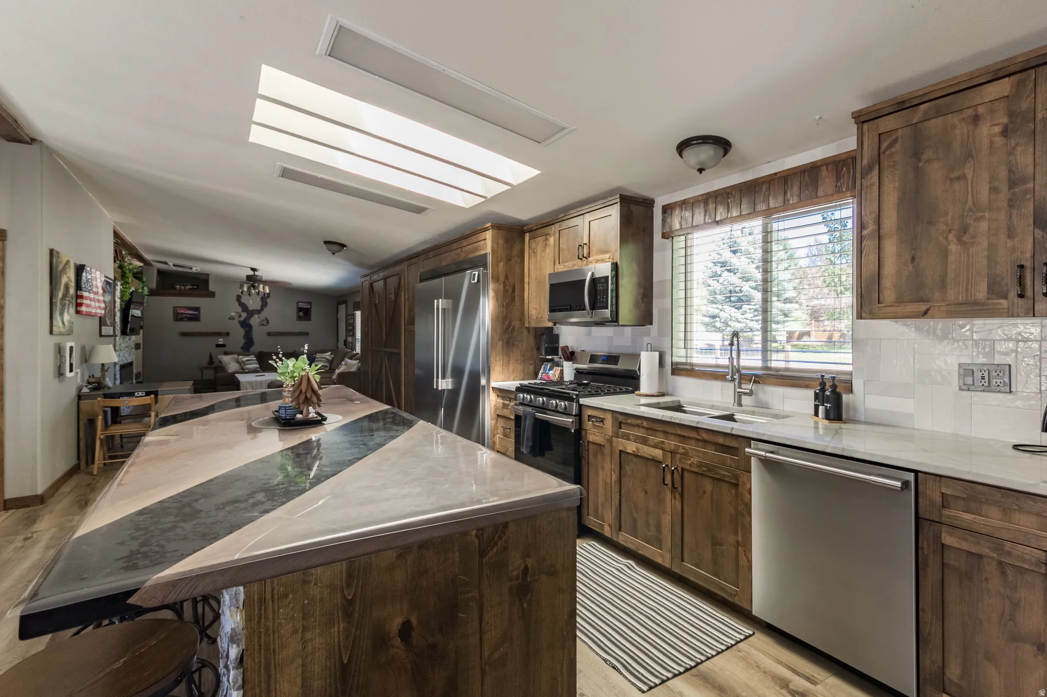 Kitchen featuring wood-finish cabinetry, stainless steel appliances, a multi-tone island countertop, a white tile backsplash, and wood-finish flooring