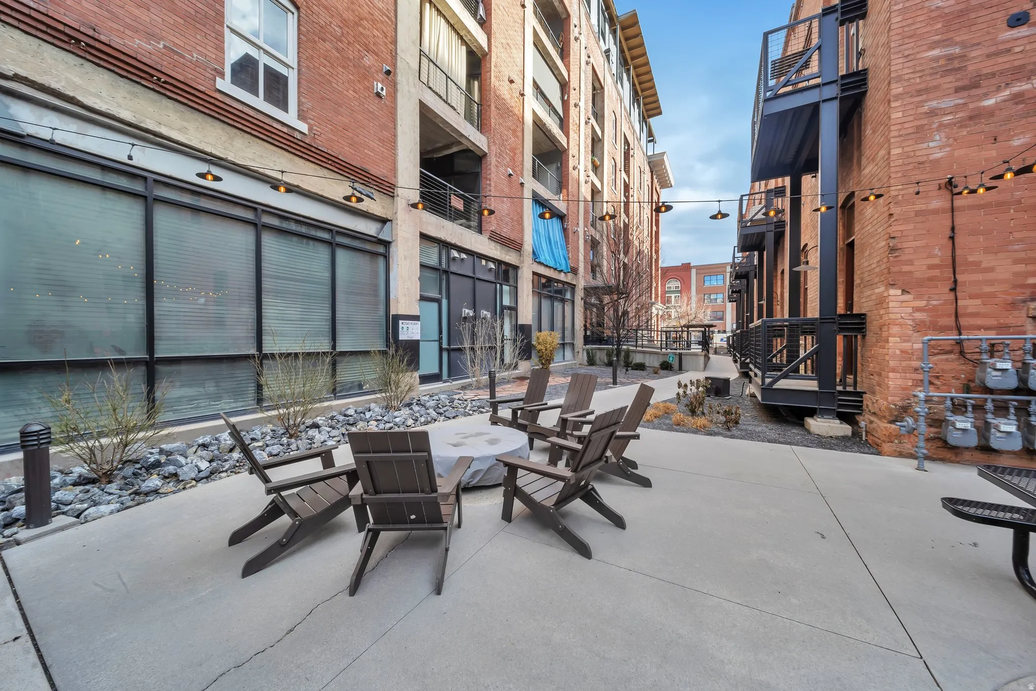 Paved courtyard featuring a central fire pit, surrounding Adirondack-style chairs, string lighting, and brick facades