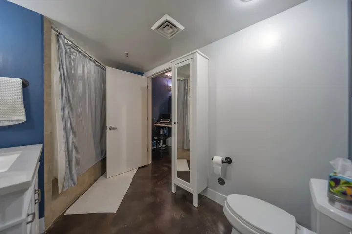 Bathroom featuring a shower with a striped curtain, a white vanity with an integrated sink, dark wood-finish flooring, a toilet, and a white storage cabinet with a mirrored door
