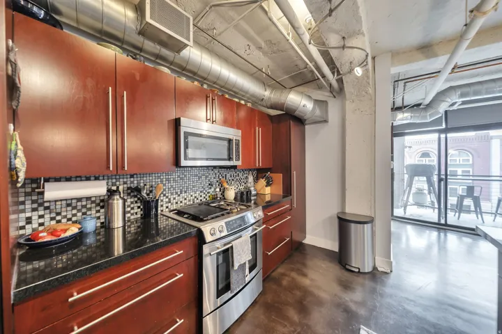 Modern kitchen featuring wood-finish cabinetry, stainless steel appliances, dark countertops, and a mosaic tile backsplash