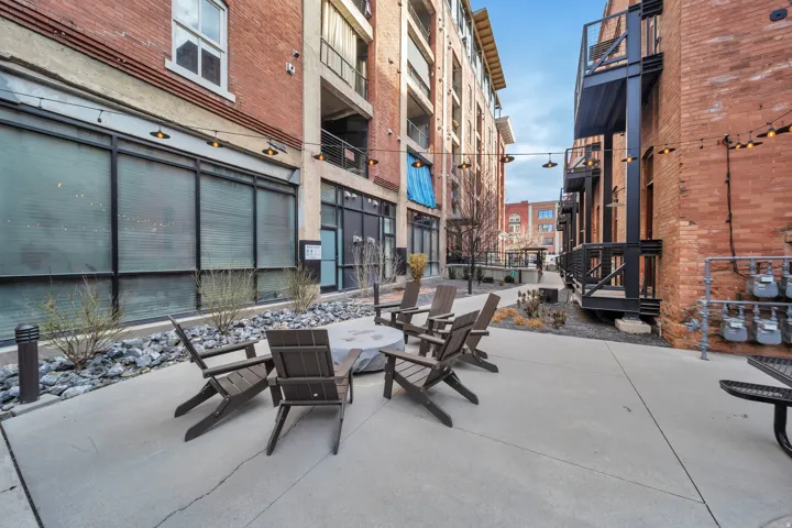 Paved courtyard featuring a central fire pit, surrounding Adirondack-style chairs, string lighting, and brick facades