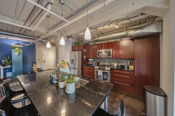 Modern kitchen featuring a large black countertop island with integrated sink, dark wood-finish cabinetry, stainless steel appliances, and a mosaic tile backsplash