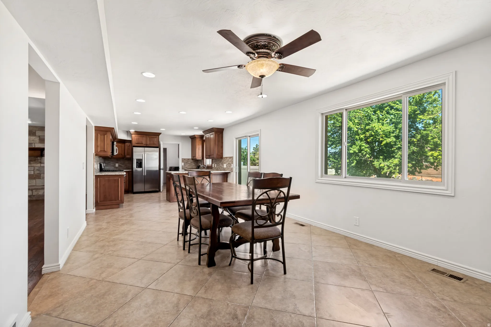 Open-concept living space featuring neutral tile flooring, recessed lighting, a ceiling fan, a large window, and wood cabinetry with stainless steel appliances