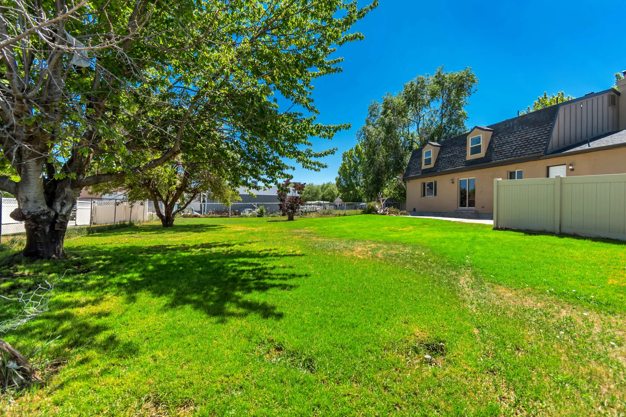Spacious green lawn with mature trees and a beige exterior structure featuring a dark shingle roof and dormer windows