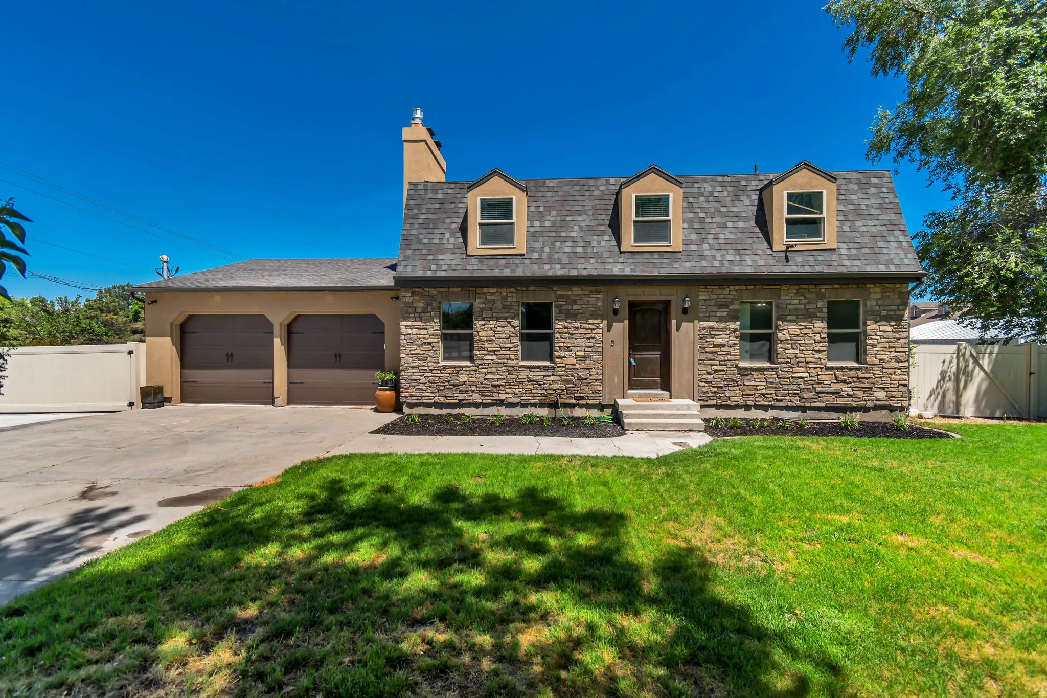 Stone facade home featuring a steep-pitched roof with three dormer windows, a two-car garage with arched entrances, a paved driveway, and an expansive front lawn