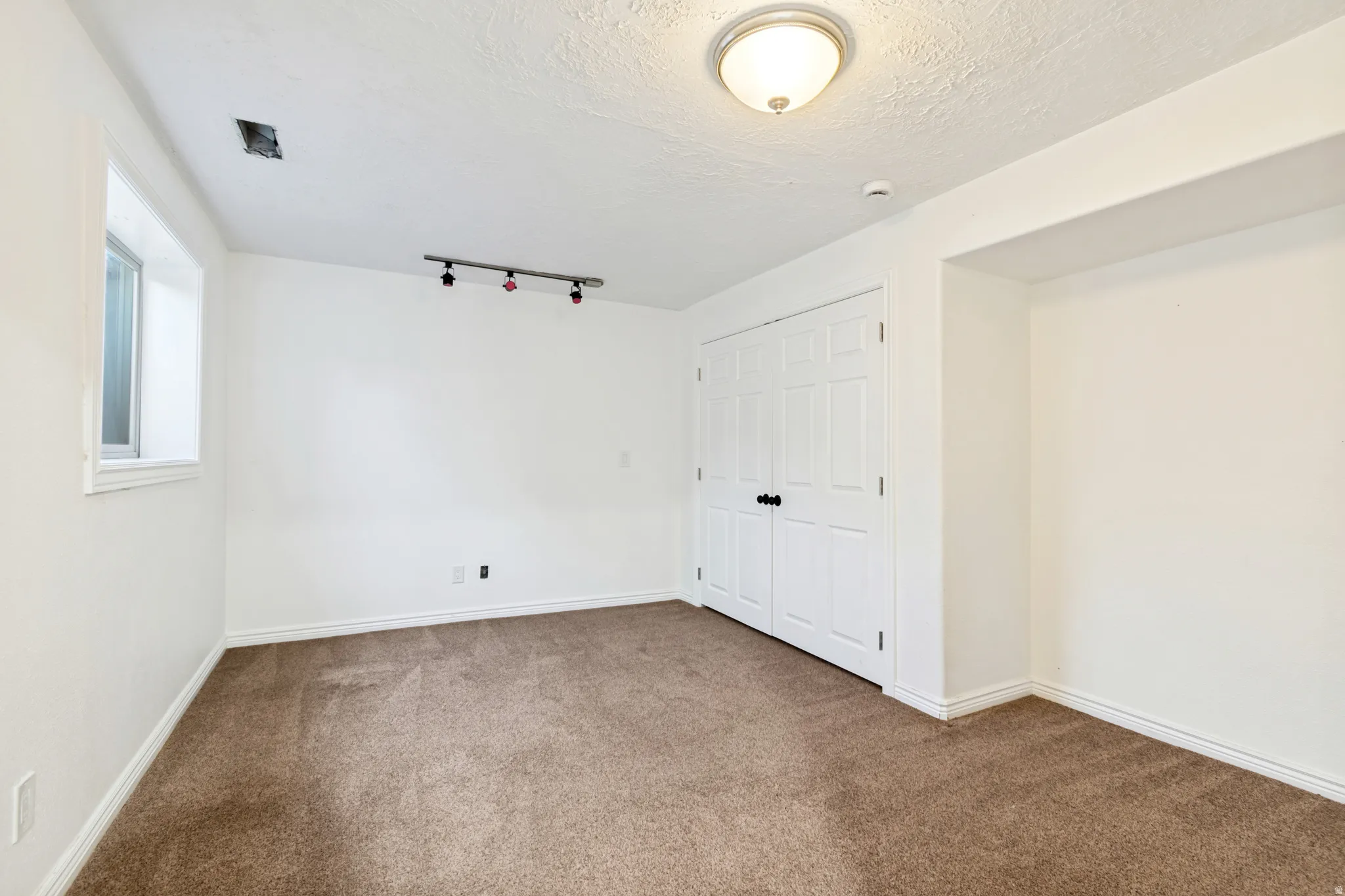 Carpeted room featuring white walls, a single window with white trim, a double bi-fold closet door with black hardware, a flush-mount ceiling light fixture, and a track lighting fixture