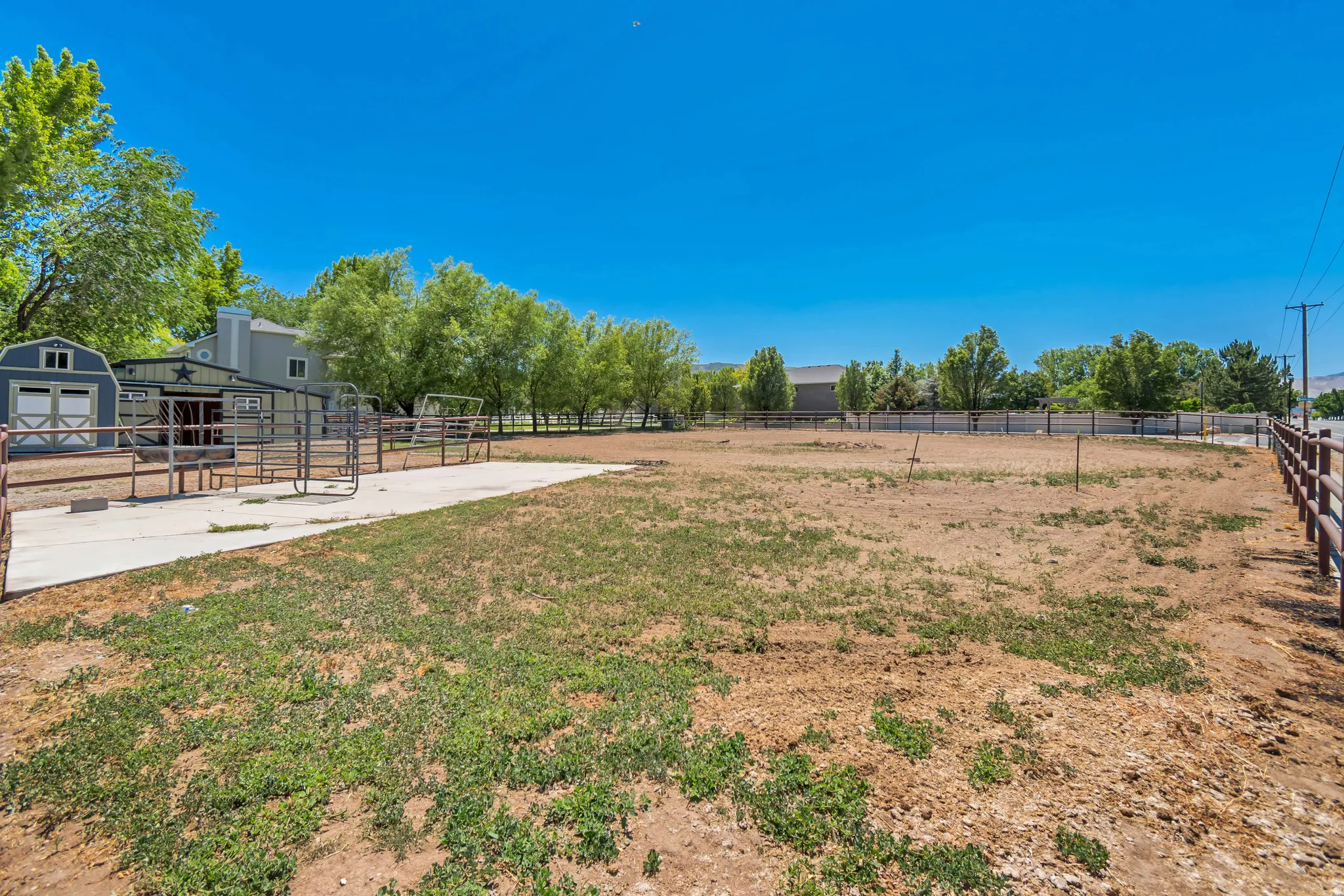 Expansive open land area featuring a large, fenced paddock, a concrete pad, a blue outbuilding with white trim, a green barn-style structure with a star emblem, and a wood-post fence