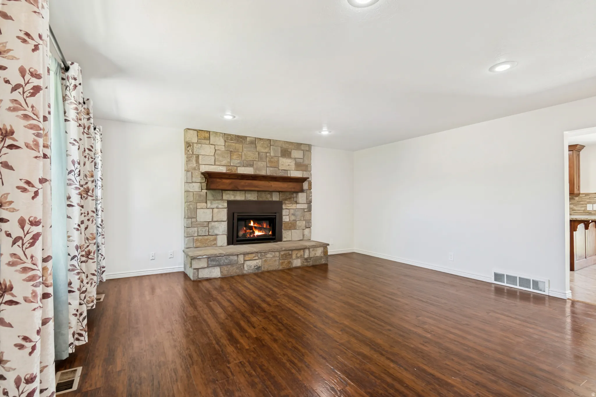 Living area featuring a stone-clad fireplace with a wood mantel, rich wood-finish flooring, recessed lighting, and a partial opening to an adjacent room with wood cabinetry and a tiled backsplash