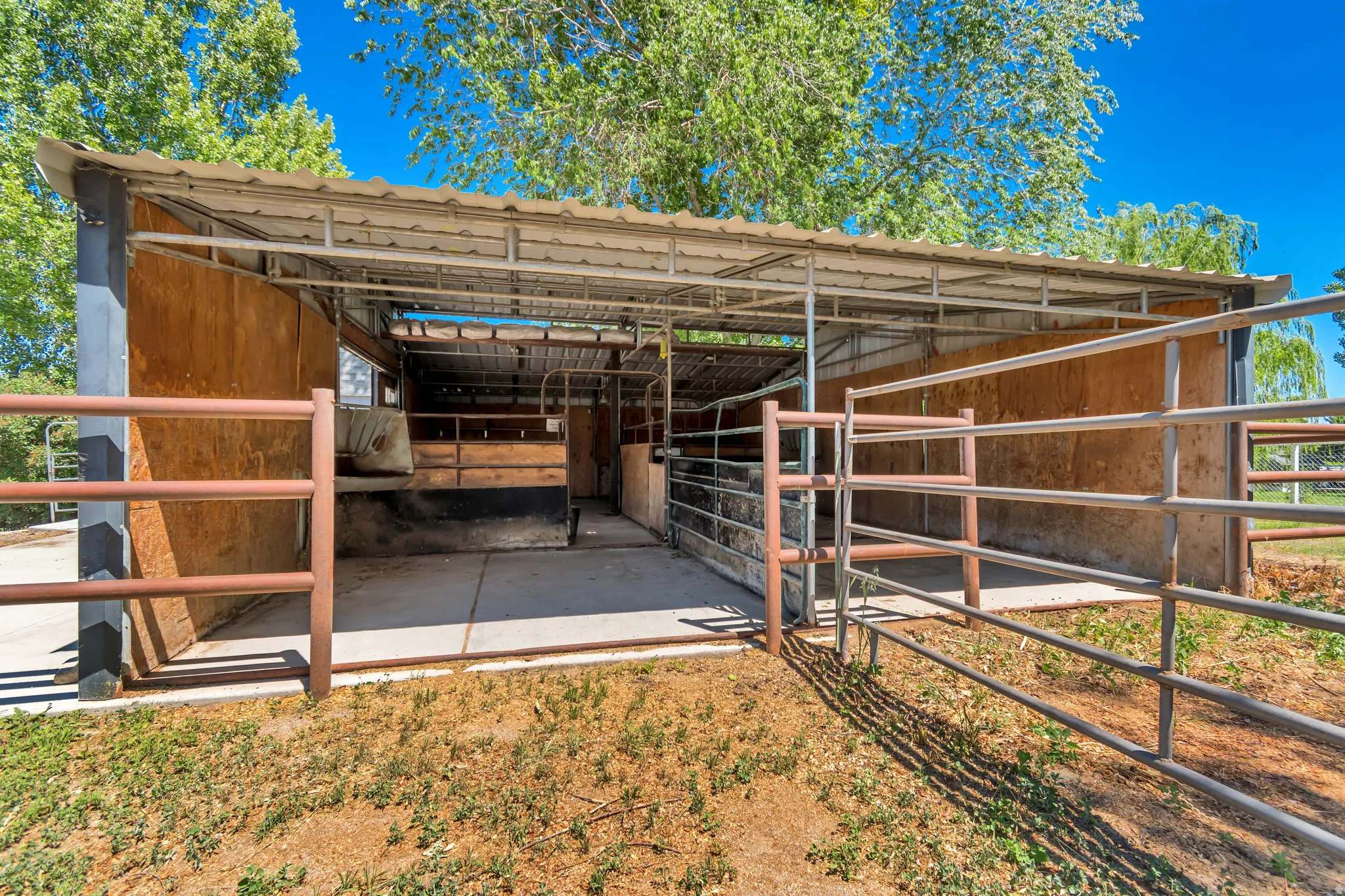 Covered stable structure featuring metal framework, a corrugated metal roof, wood-paneled stalls, and metal fencing