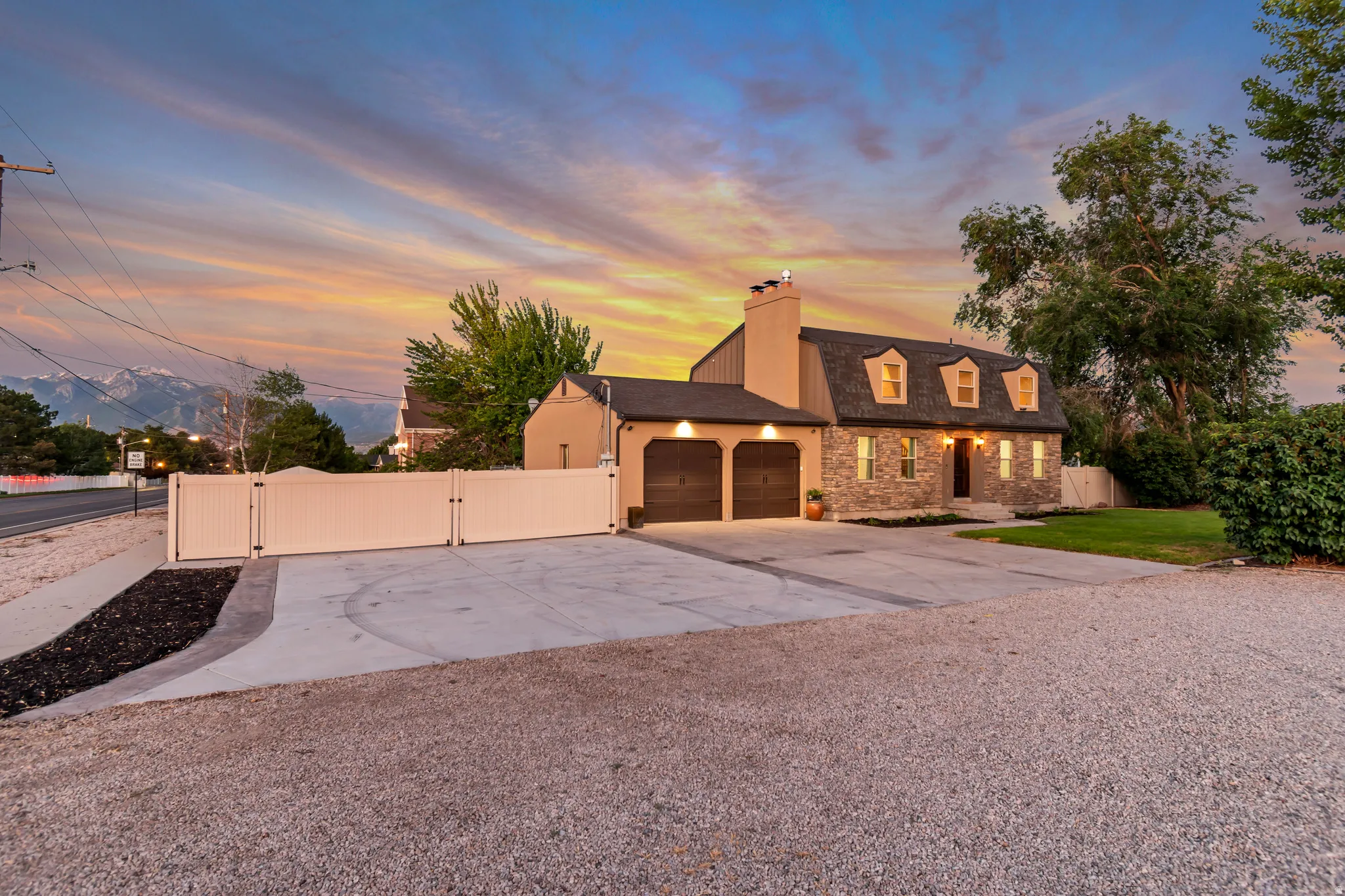 Stone and stucco exterior with dormer windows, featuring a two-car garage with wood-finish doors, a paved driveway, and a white privacy fence