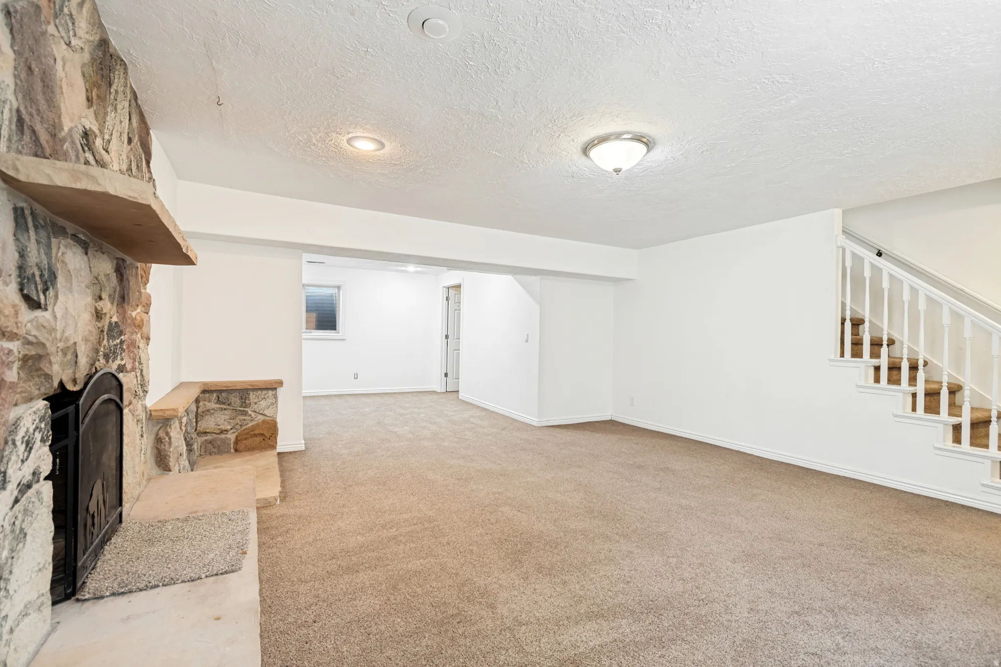Expansive carpeted room featuring a stone-clad fireplace with a hearth and mantel, white walls, recessed lighting, and a staircase with white balusters