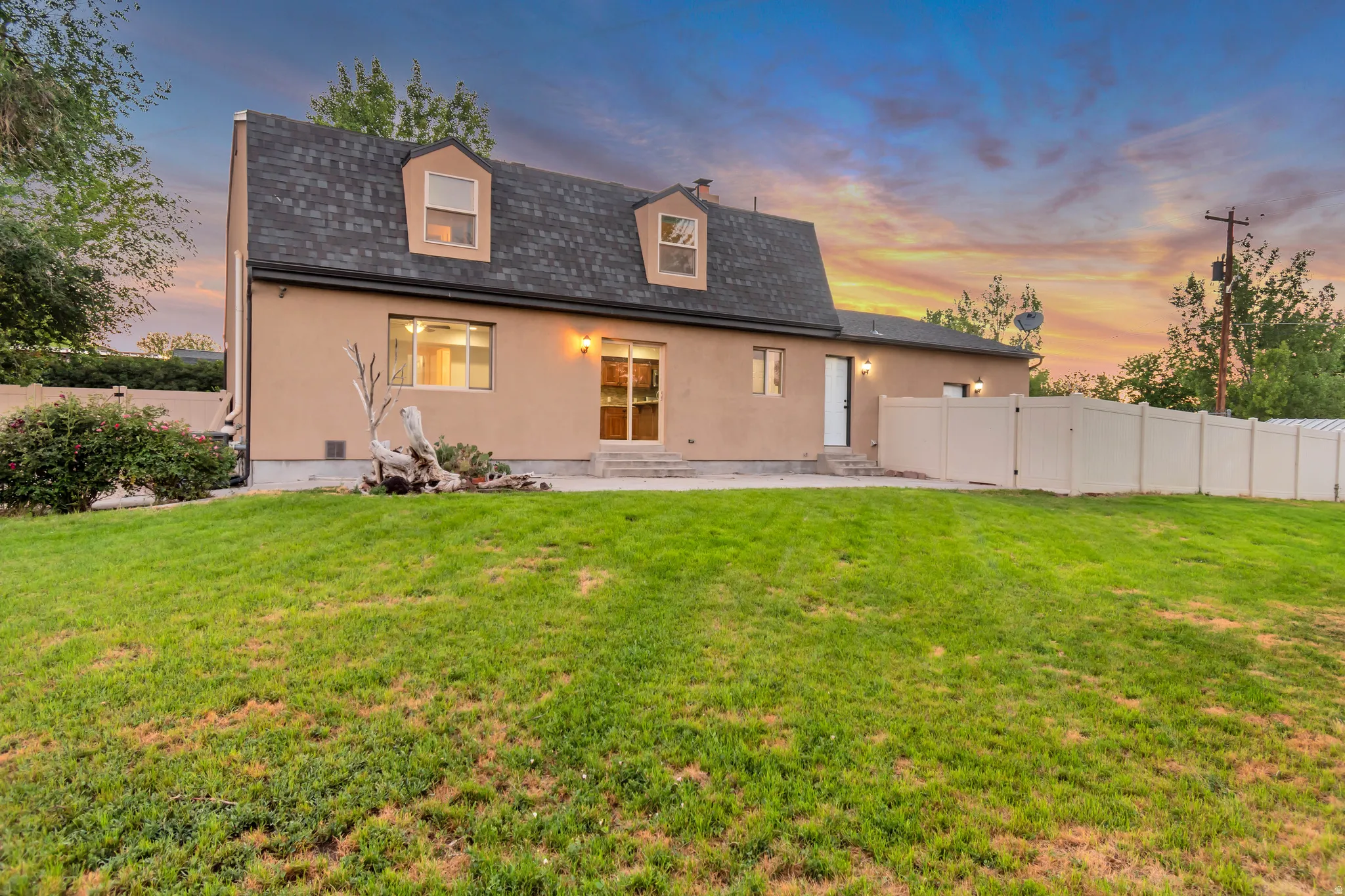 Spacious rear yard with a sloped lawn, stucco exterior, dormer windows, sliding glass door access, and privacy fencing