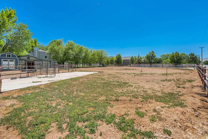 Expansive open land area featuring a large, fenced paddock, a concrete pad, a blue outbuilding with white trim, a green barn-style structure with a star emblem, and a wood-post fence