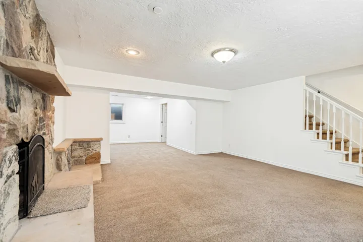 Expansive carpeted room featuring a stone-clad fireplace with a hearth and mantel, white walls, recessed lighting, and a staircase with white balusters