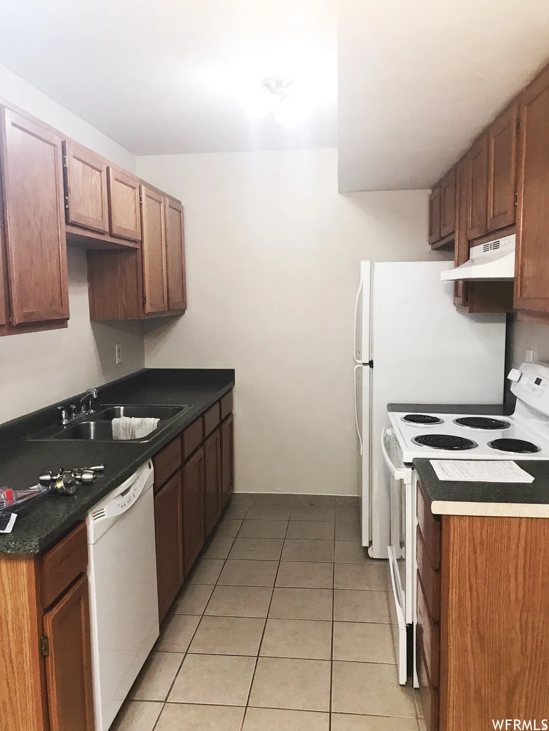 Kitchen featuring wood cabinetry, dark countertops, a double-basin stainless steel sink, white dishwasher, white electric range, and a white refrigerator