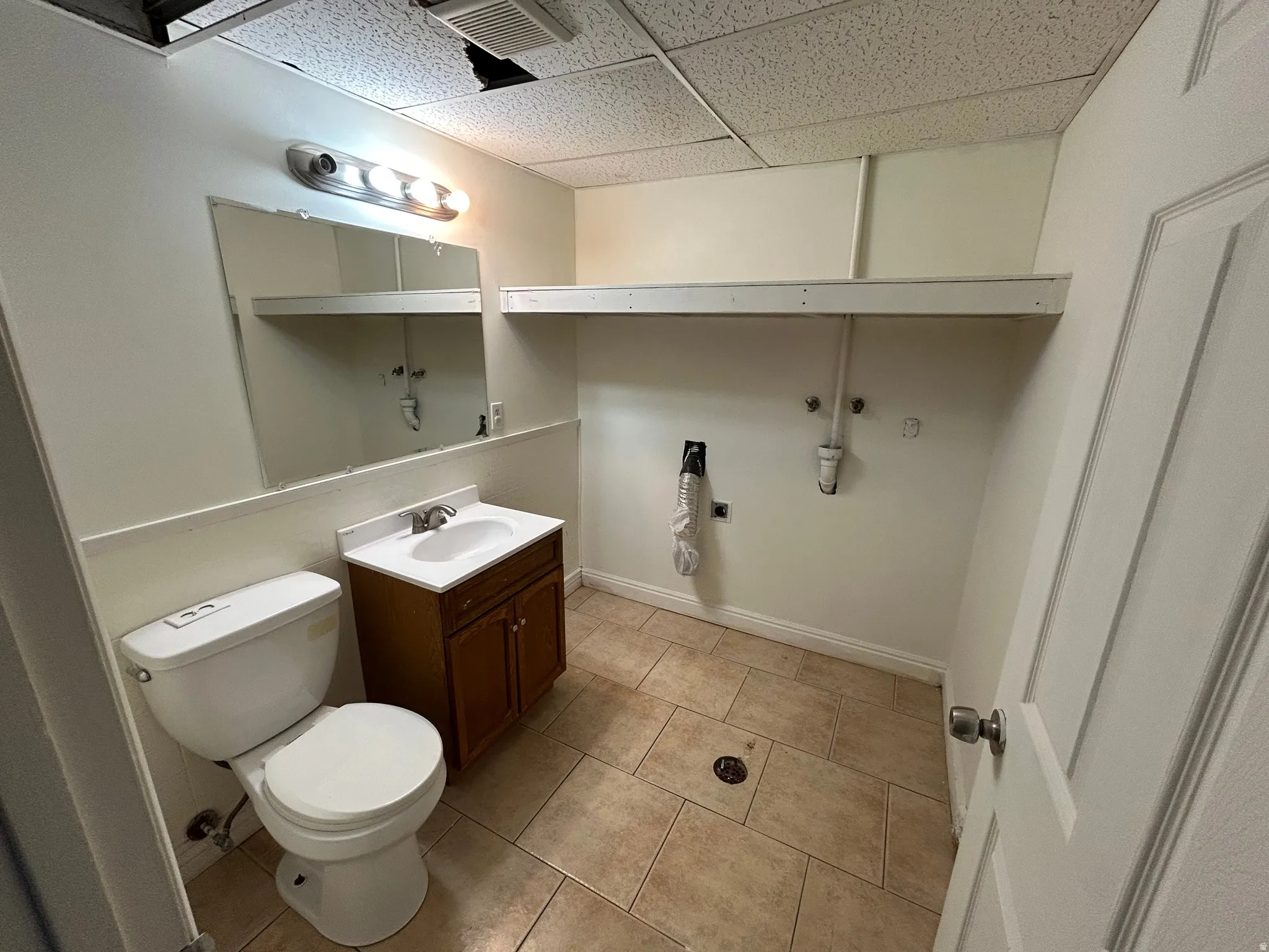 Functional bathroom featuring a drop ceiling, tile flooring, a single vanity with a white countertop, and a wall-mounted mirror with an overhead light fixture