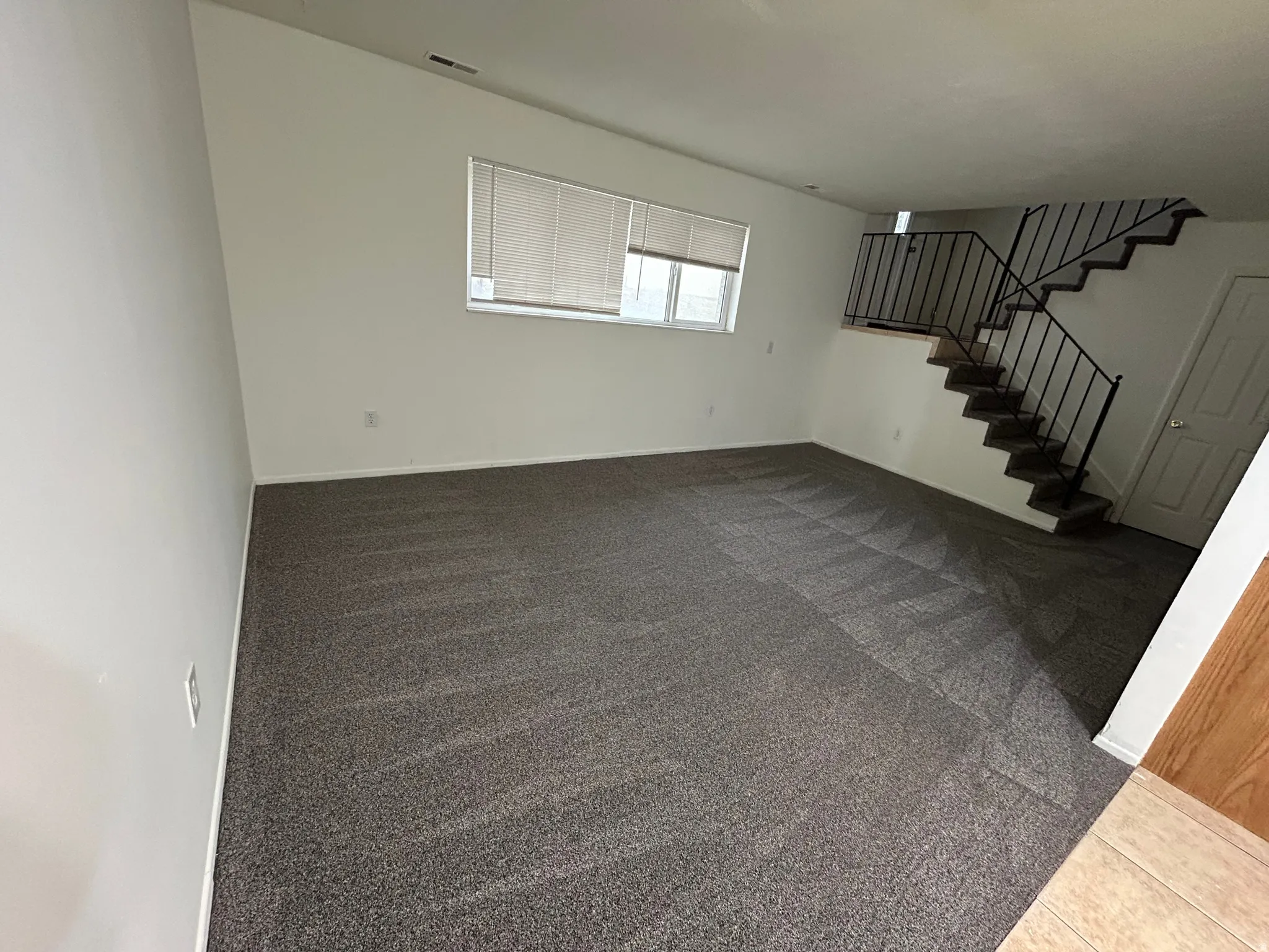 Spacious living area featuring neutral carpeting, white walls, and a large window with blinds
