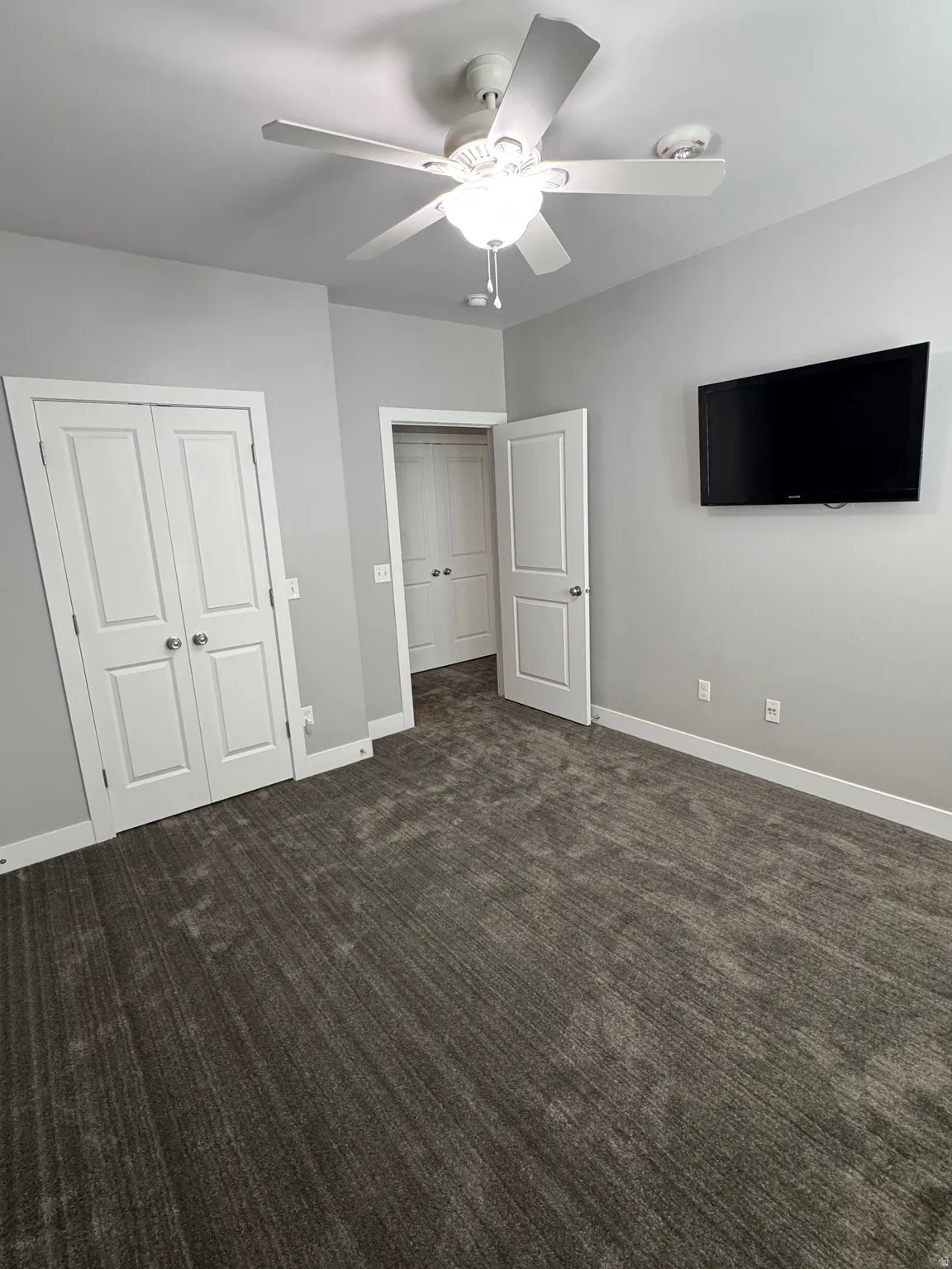 Spacious room featuring neutral gray walls, charcoal-toned carpeting, white trim, and a ceiling fan with integrated lighting