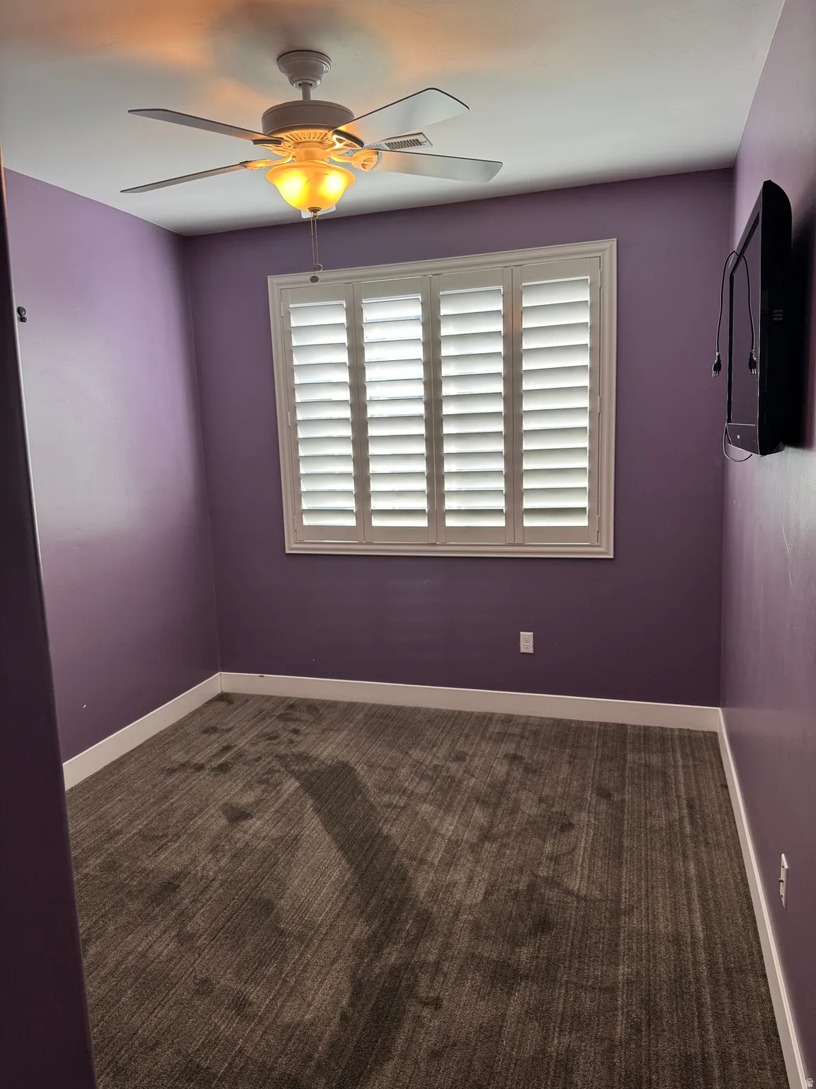 Room featuring a ceiling fan with integrated lighting, window with white plantation shutters, rich purple wall color, and striped carpet flooring