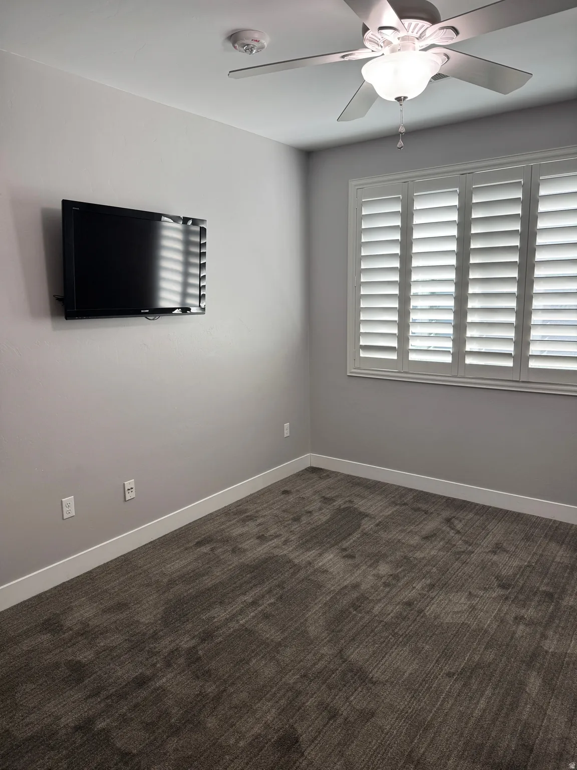 Room featuring a ceiling fan with integrated lighting, window with plantation shutters, and textured gray carpeting