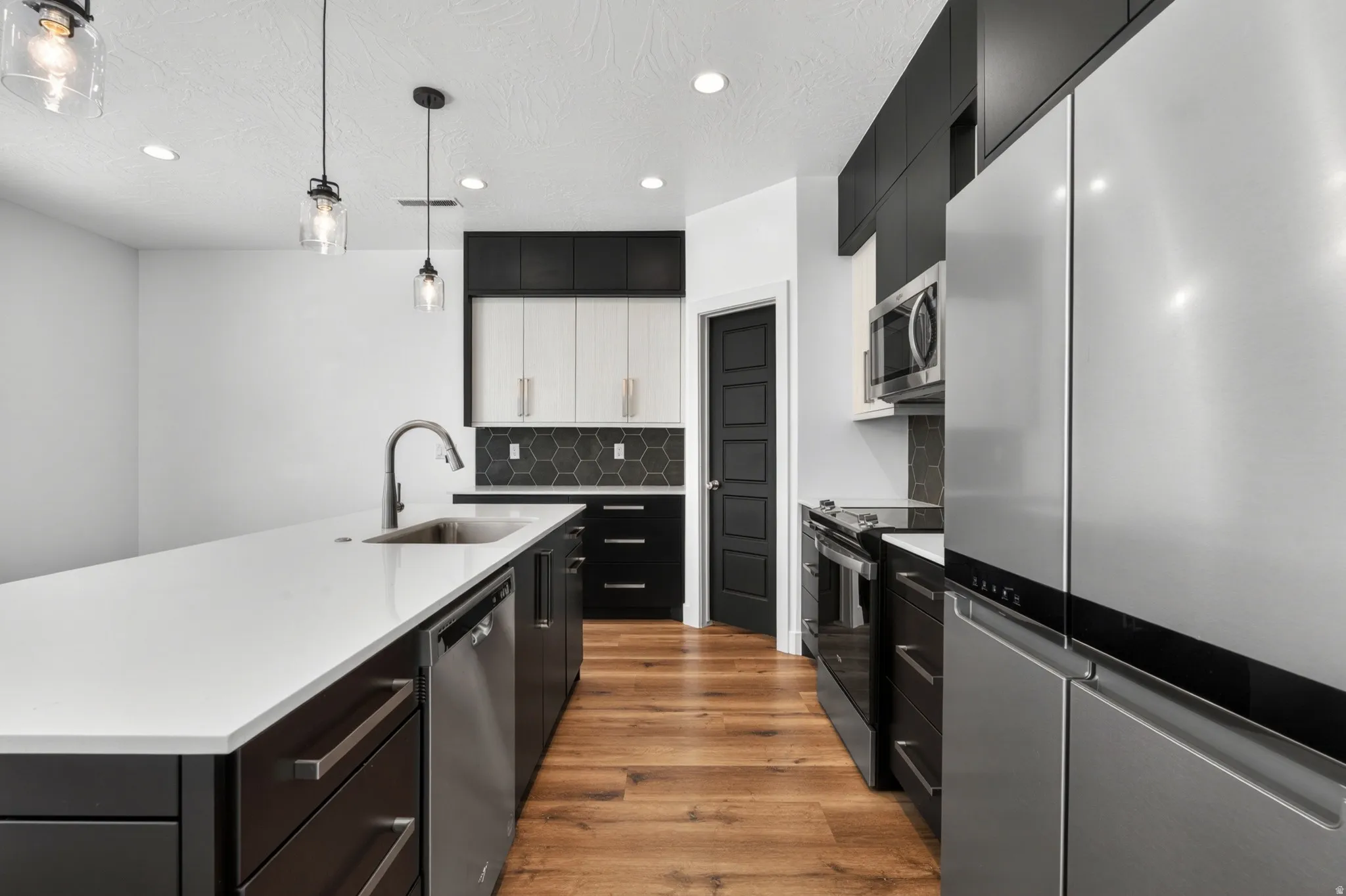 Modern kitchen featuring a large island with an integrated sink, stainless steel appliances, dark cabinetry, wood-finish flooring, and pendant lighting fixtures