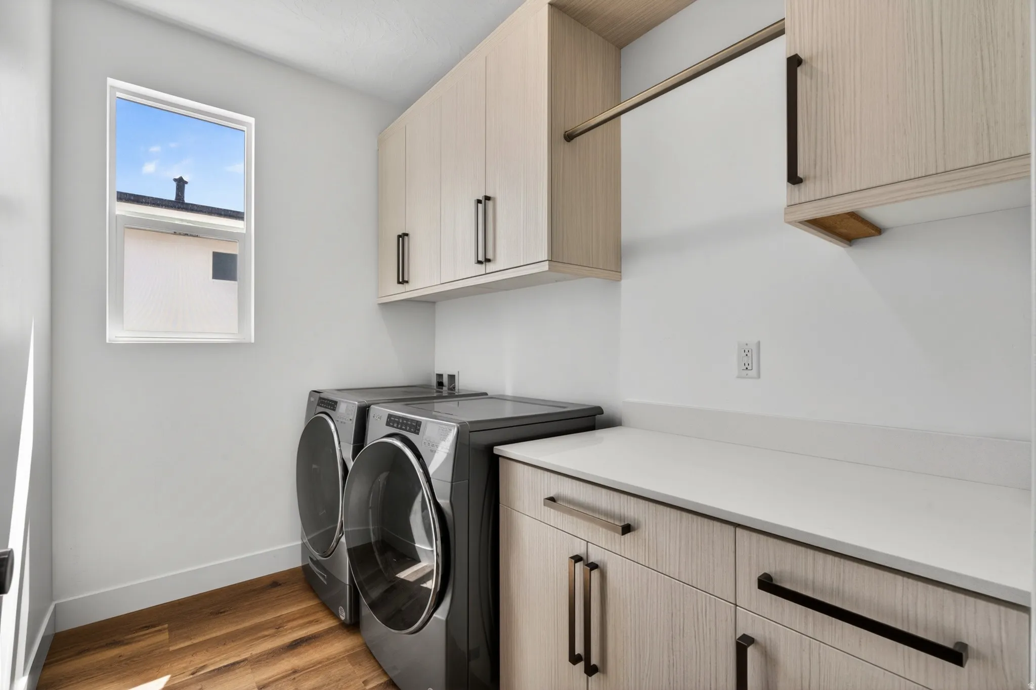 Laundry area featuring wood-finish flooring, a white-framed window, light-toned cabinetry with dark hardware, a white countertop, and a built-in hanging rod