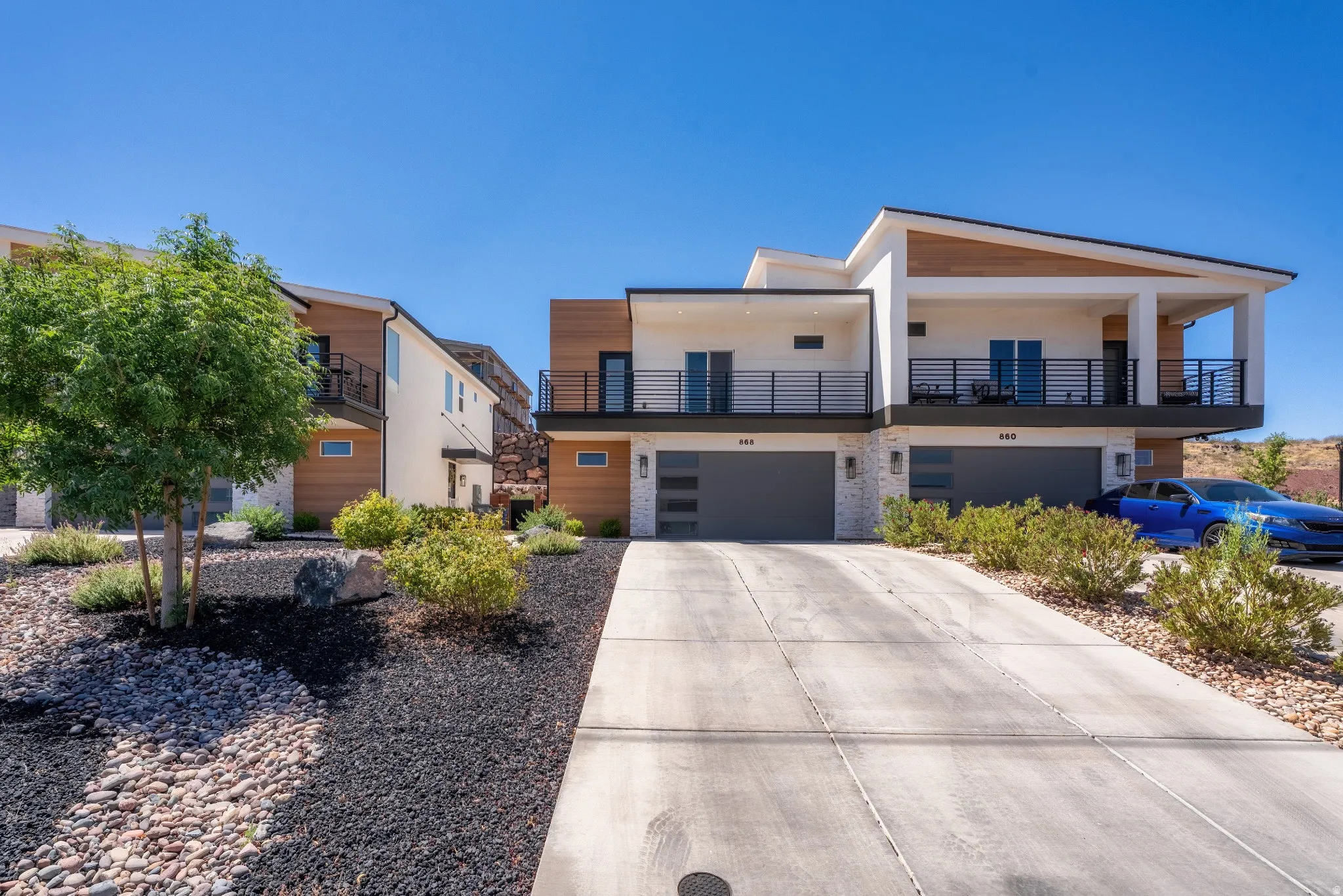 Modern desert-style exterior featuring a cantilevered balcony, integrated garages with opaque glass panels, concrete driveways, and low-maintenance xeriscaping with dark mulch and natural stone accents