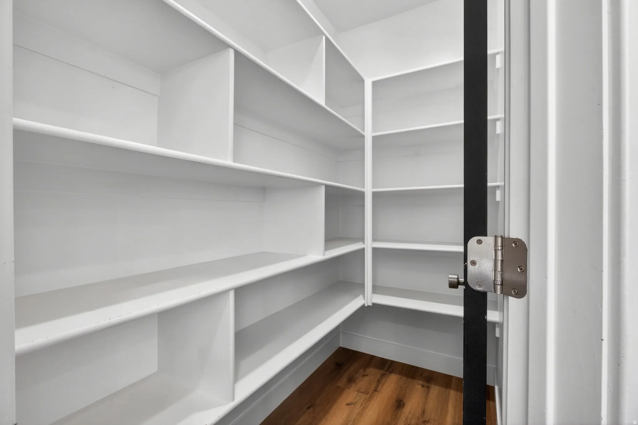 Walk-in pantry featuring extensive white shelving, wood-finish flooring, and a white door with a black frame