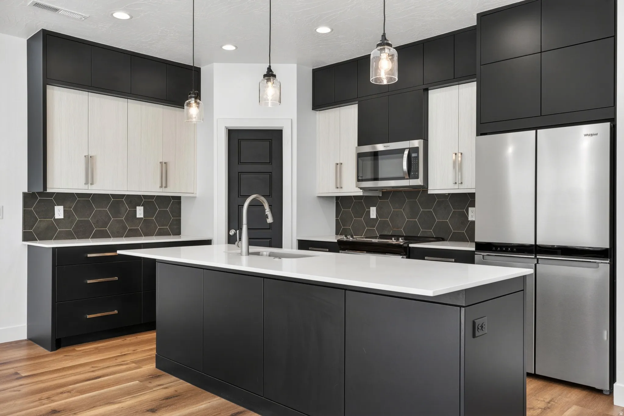 Modern kitchen featuring a large central island with a white countertop, two-tone cabinetry in black and light wood-finish, stainless steel appliances, and a hexagonal tile backsplash
