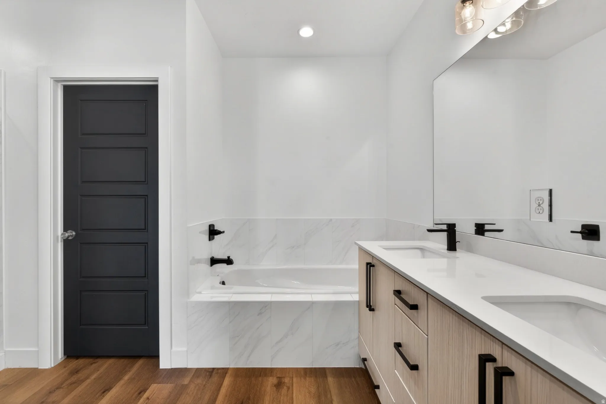 Bathroom featuring a wood-finish double vanity with white countertops and undermount sinks, black hardware and faucets, an integrated bathtub with white tile surround, and wood-finish flooring