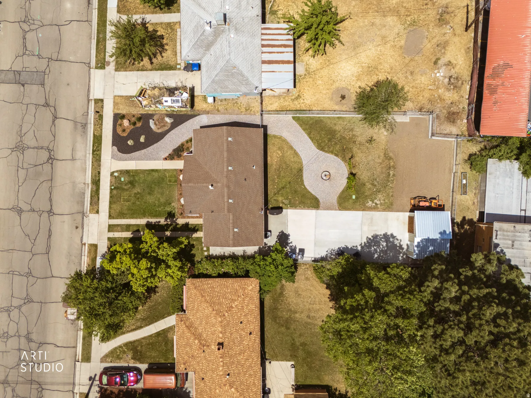 Aerial property overview featuring a brown shingle roof, a concrete driveway, a paved pathway with a circular patio, a gravel area, and mature trees