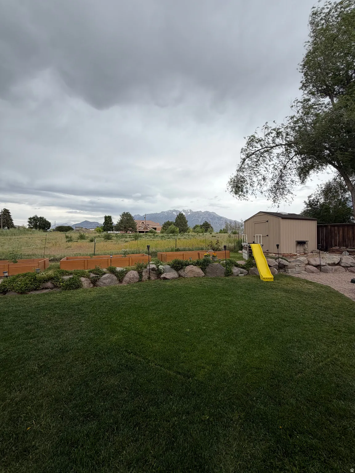 Expansive backyard featuring a manicured lawn, established garden beds with wood-finish frames, a rock retaining wall, and a storage shed