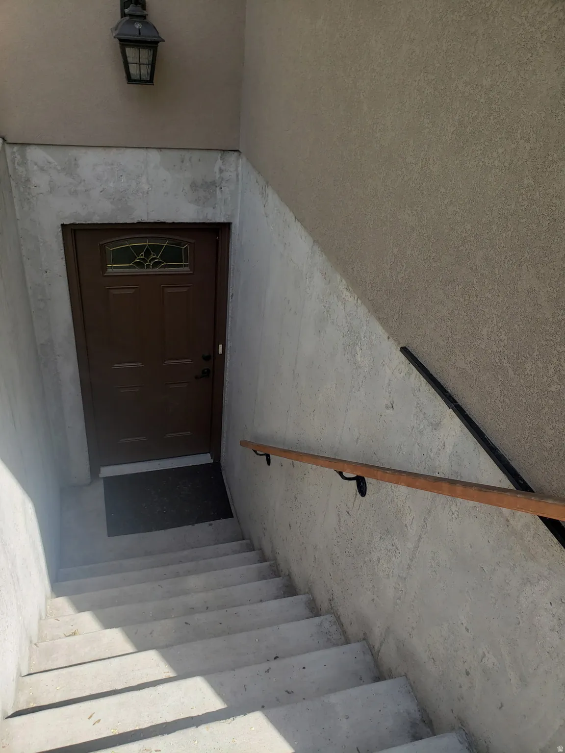 Entryway featuring a wood-panel door with decorative glass, stucco exterior, mounted outdoor lantern, concrete stairs, and a wood handrail with metal brackets