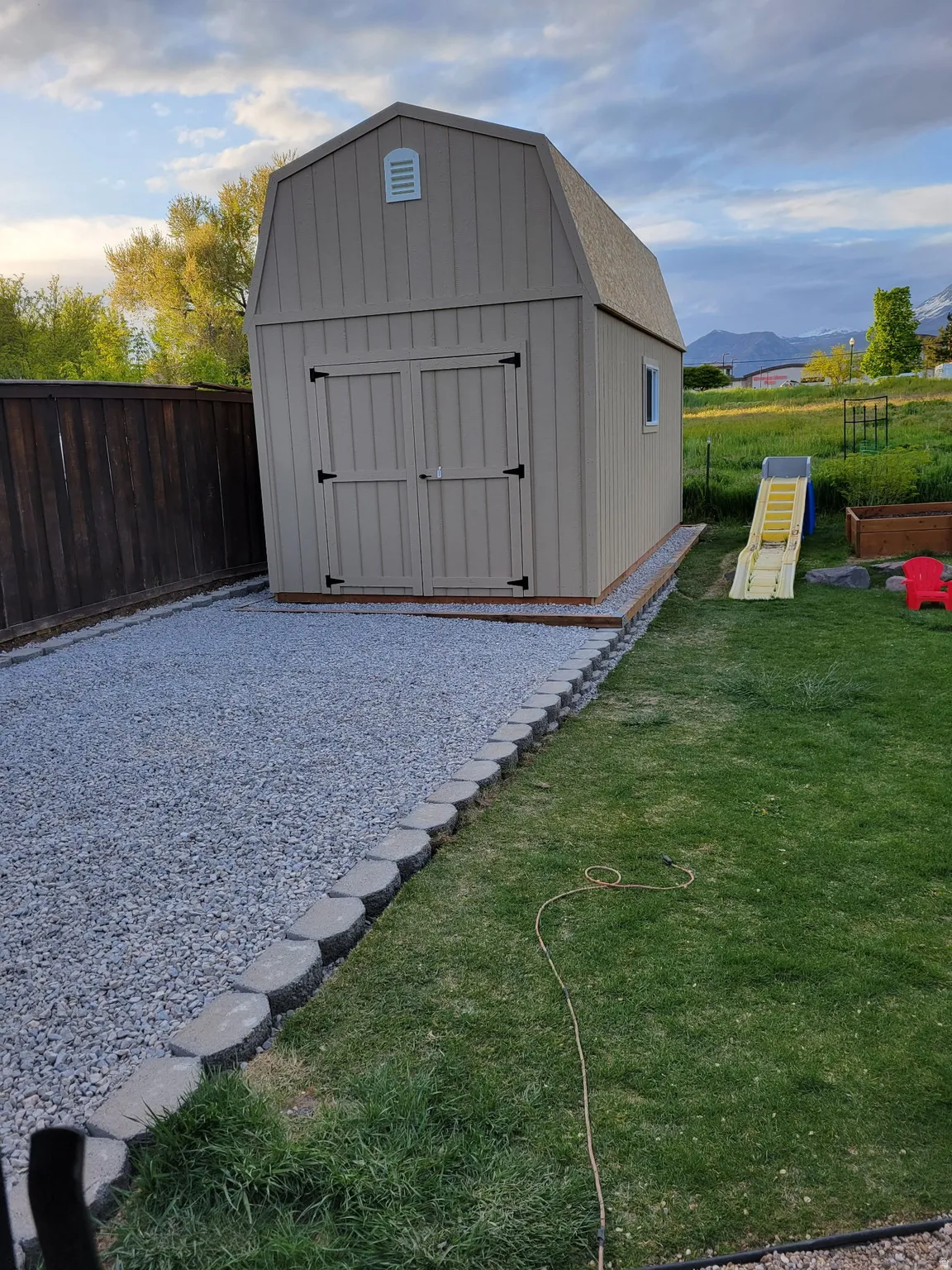 Gambrel-style storage shed with double doors and a side window, framed by a gravel path and a wood fence