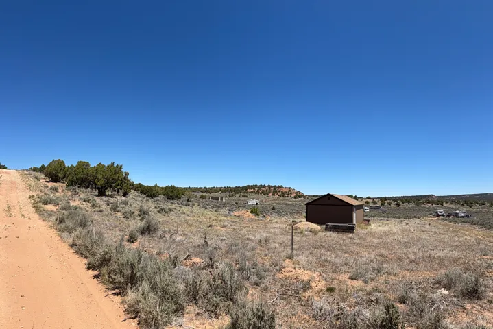 Expansive open land featuring a brown outbuilding, an unpaved road, and distant hillside vegetation