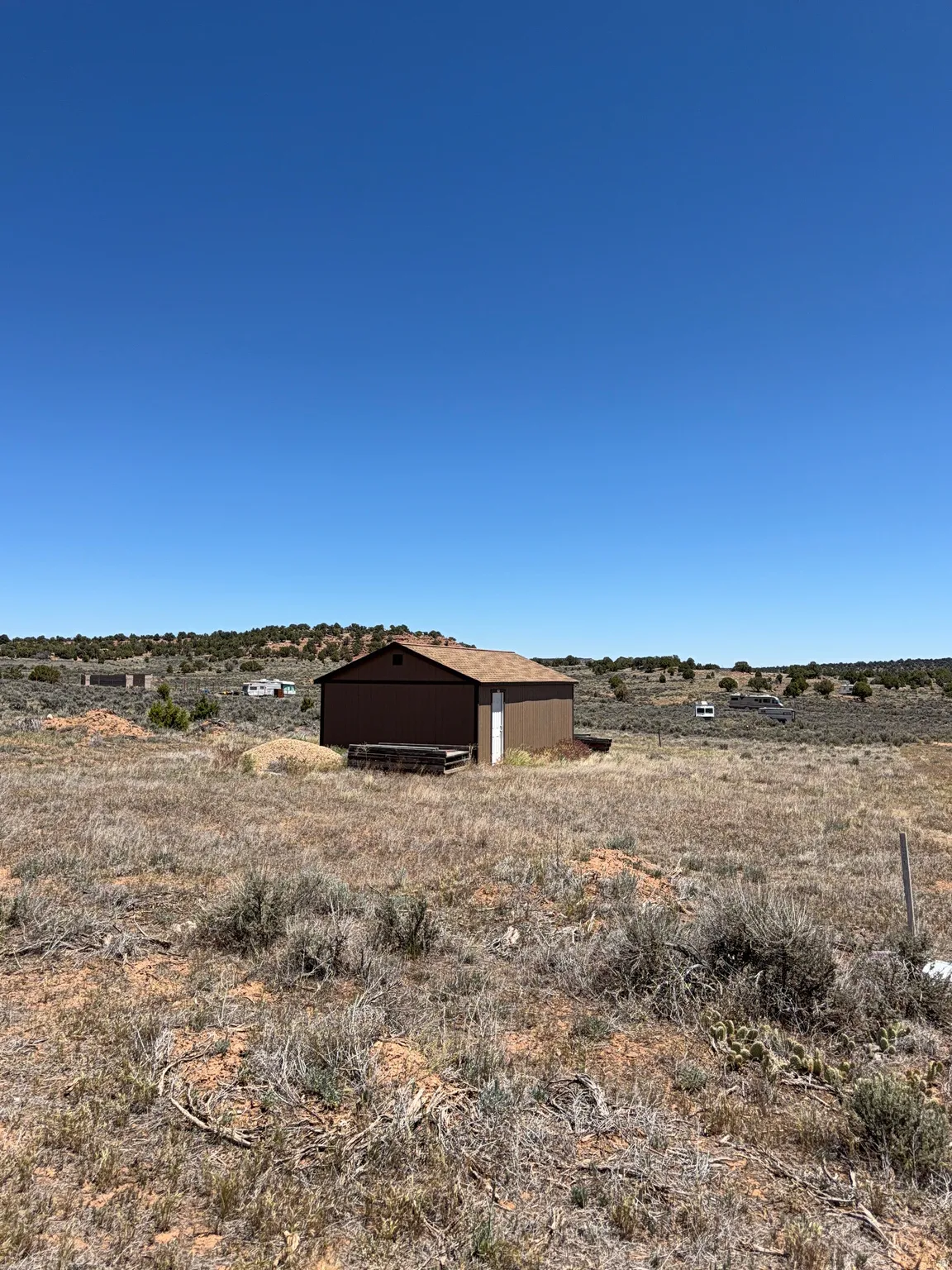 Brown metal outbuilding with a single white door and a brown shingle roof, set on a natural ground surface with sparse vegetation