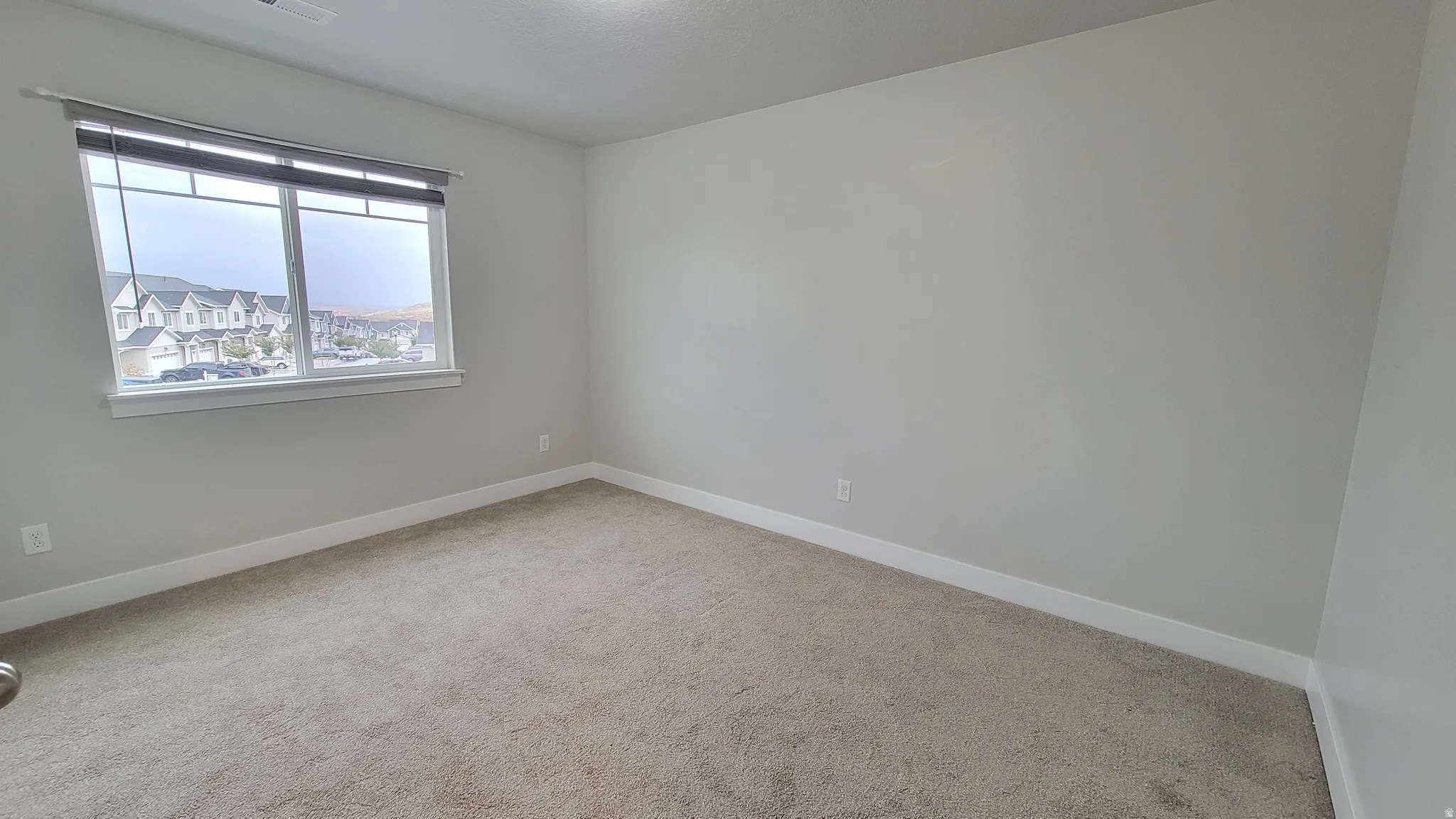 Carpeted room featuring a large window with roll-up blinds, neutral paint tones, and white baseboards