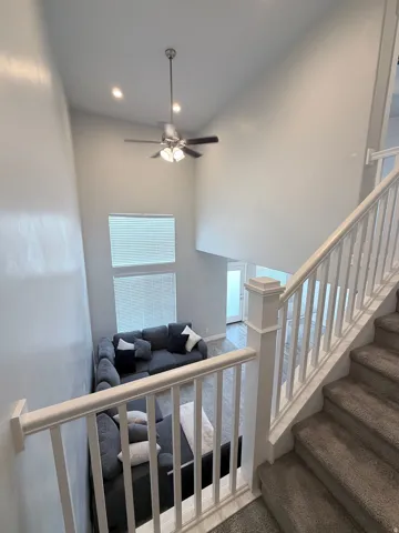 Staircase with carpeted treads and white wood balustrade overlooking a living area with high ceilings, recessed lighting, and a ceiling fan