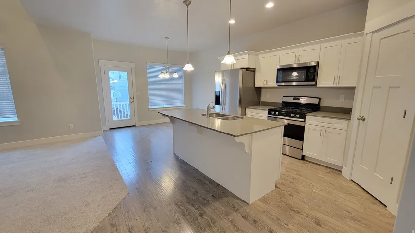 Open-concept kitchen featuring white cabinetry, stainless steel appliances, a central island with a sink, pendant lighting, and wood-finish flooring