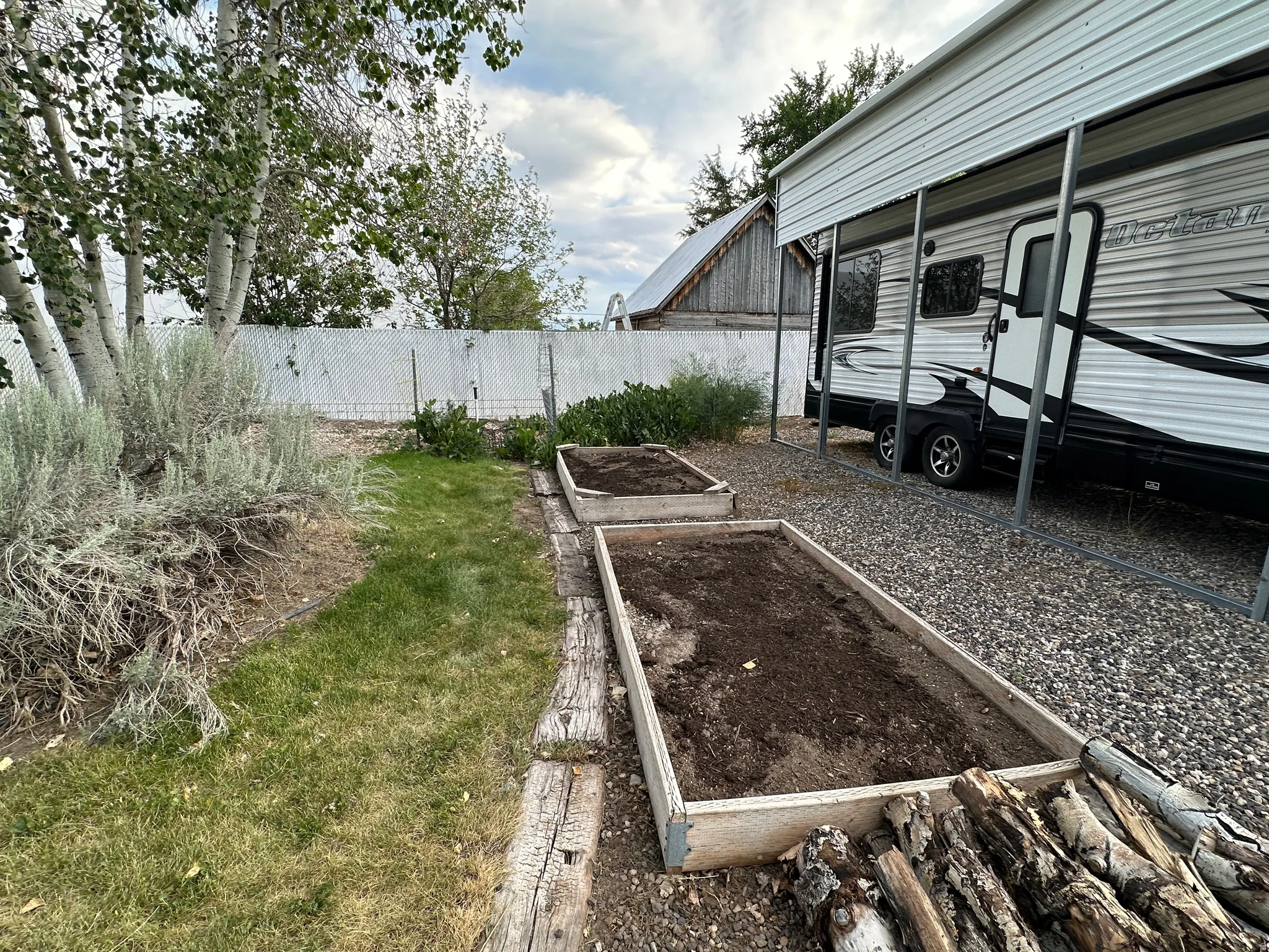 Gravel parking area featuring a metal carport structure, two raised garden beds, and a white chain-link fence