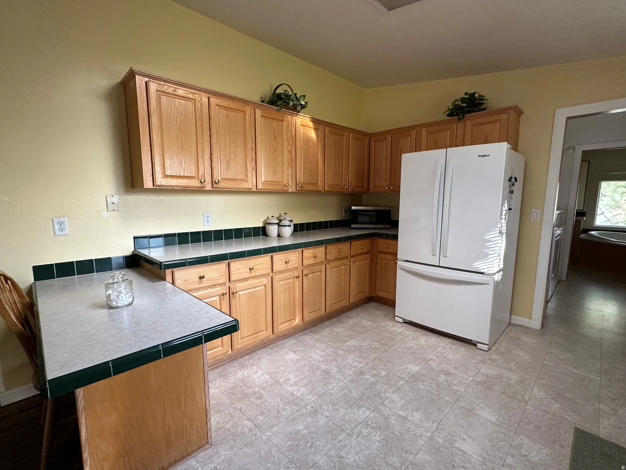 Kitchen featuring wood cabinetry, light-toned countertops with dark green tile trim, a white French door refrigerator, and light gray tile flooring