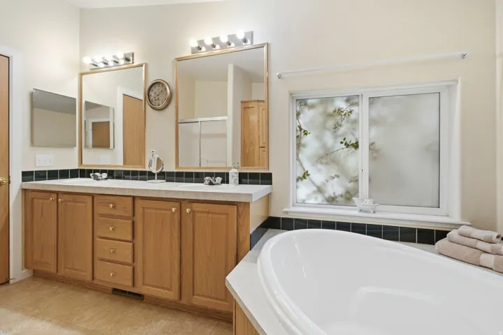 Spacious bathroom featuring a dual vanity with light wood cabinetry and integrated sinks, a large soaking tub, and a window with obscured glass